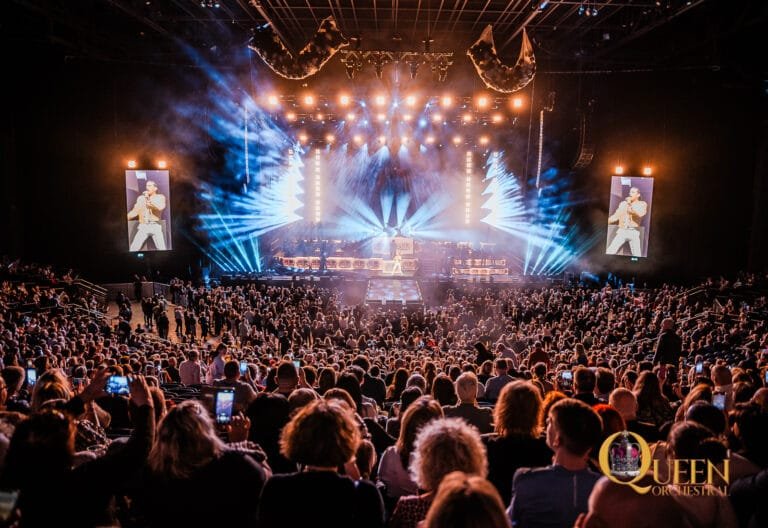 Wide-angle shot of a lit-up arena stage during QUEEN Orchestral's live show at 3 Arena, with dramatic spotlights cutting through haze and a sold out arena audience.