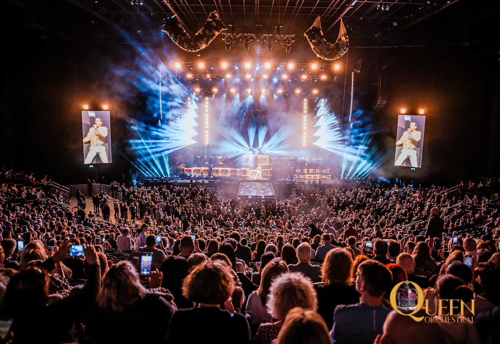 Wide-angle shot of a lit-up arena stage during QUEEN Orchestral's live show at 3 Arena, with dramatic spotlights cutting through haze and a sold out arena audience.